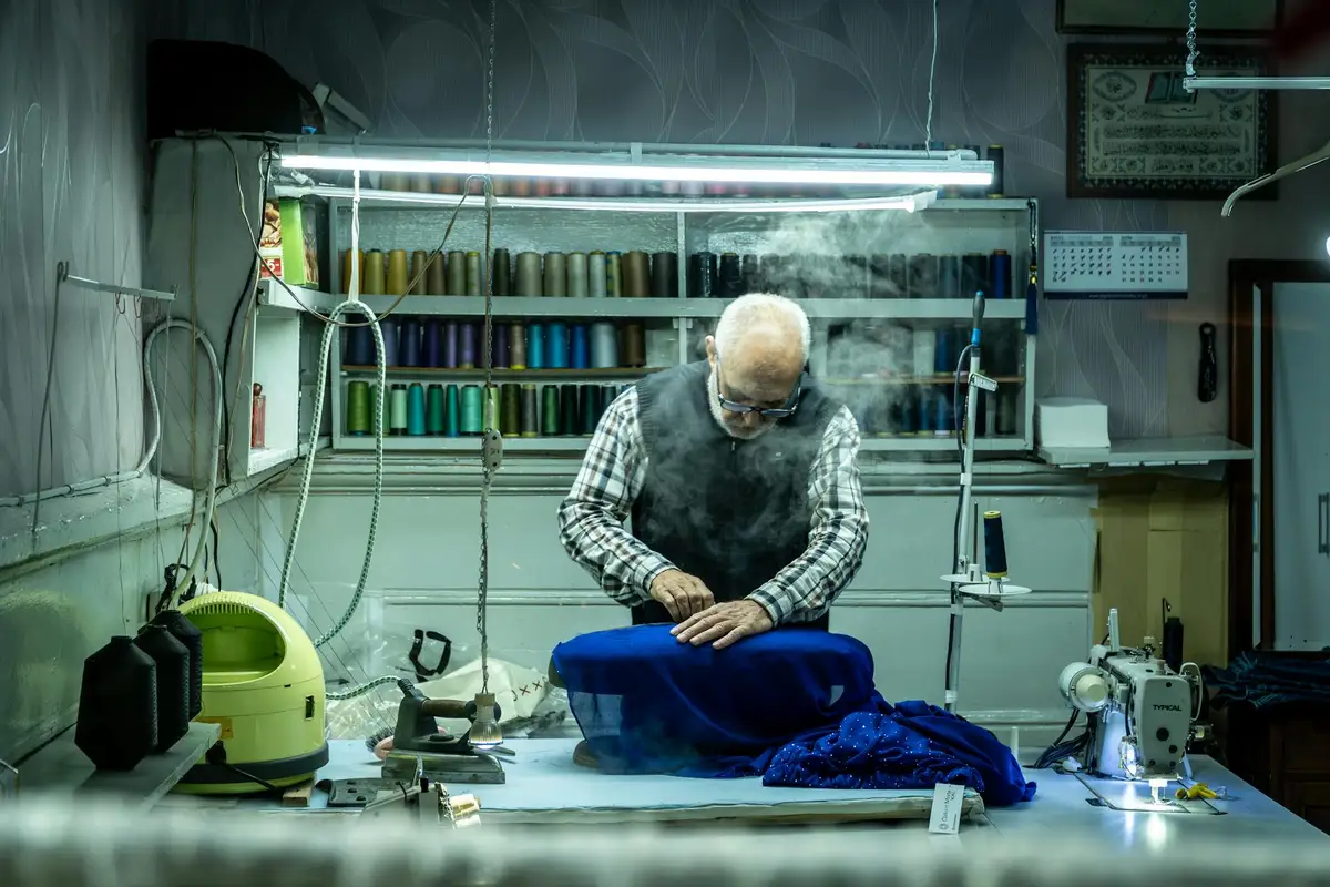 An artisan in a workshop presses a blue linen fabric on a worktable, with a bright overhead light and sewing supplies in the background.