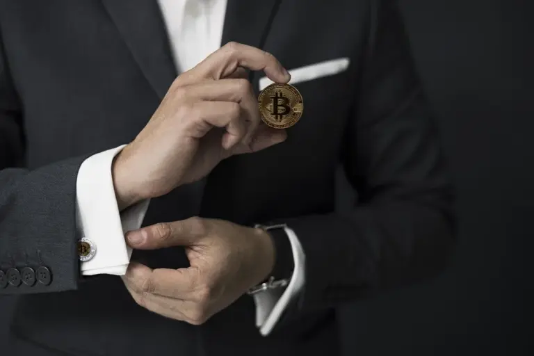A person in a dark suit adjusts cufflinks while holding a Bitcoin coin between their fingers.