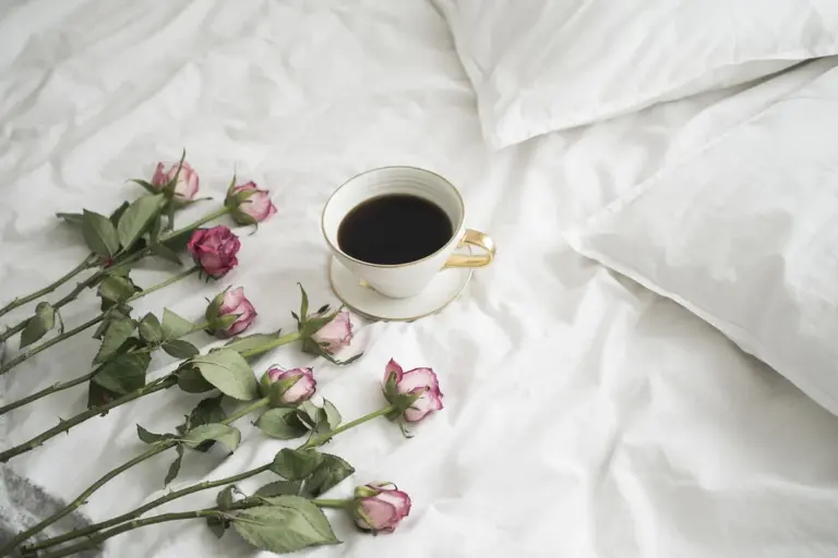White linen sheets with a cup of coffee and pink roses laid on a bed.