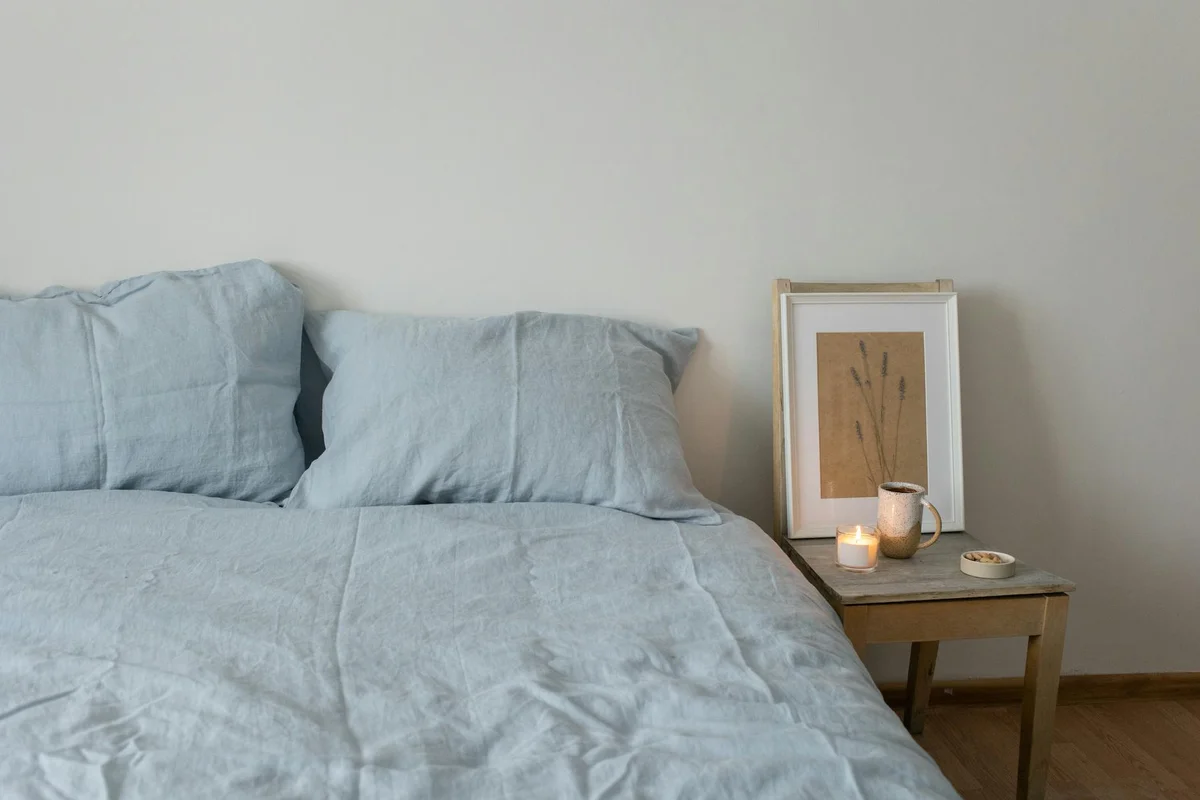 A neatly made bed with light blue linen bedding and two pillows, beside a wooden nightstand with a framed artwork, candles, and small decor.