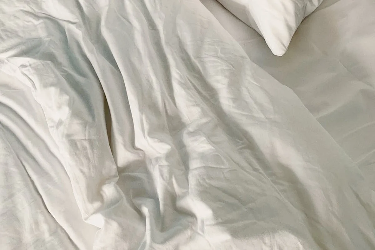 White linen sheets with natural wrinkles spread over a bed, with a pillow in the corner.