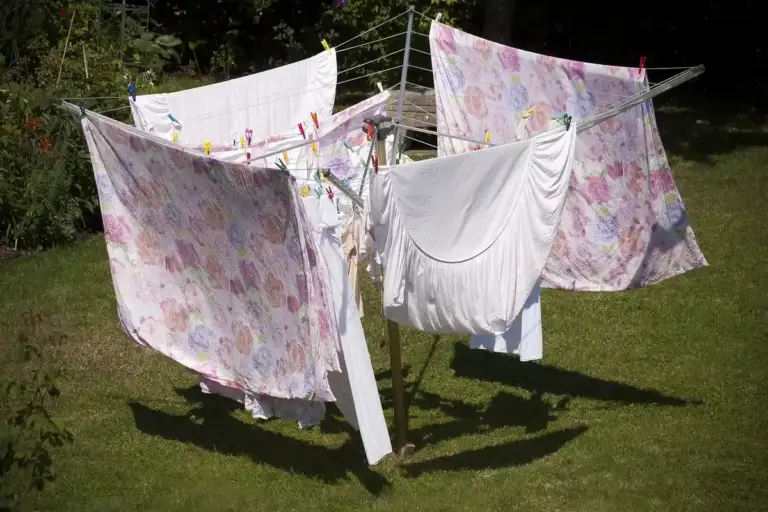 Linen and cotton fabrics drying on a clothesline in a sunny backyard, with floral-pattern sheets and plain white garments.