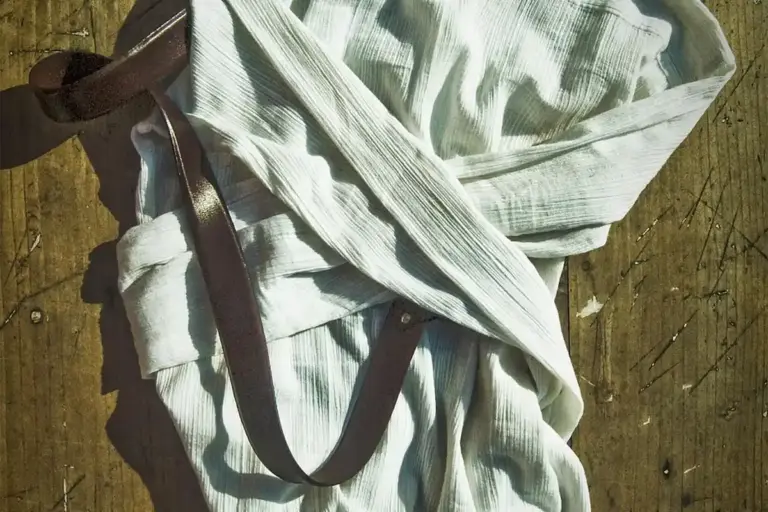 Light, wrinkled linen fabric with a brown leather strap laid on a wooden surface
