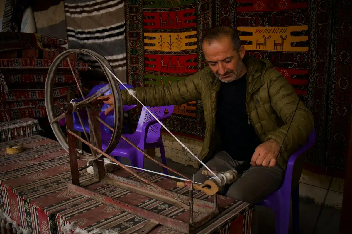 Man weaving on a loom in a workshop with colorful woven rugs hanging on the wall in the background