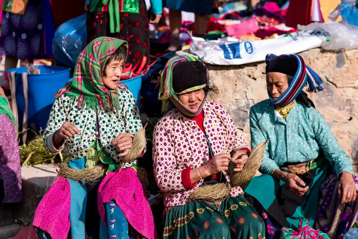 Three women in bright, patterned traditional clothing sit at a market stall weaving fabric and sorting threads.