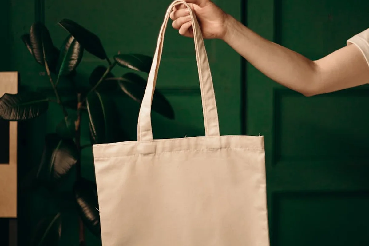 Beige tote bag suspended by its strap against a dark green background.