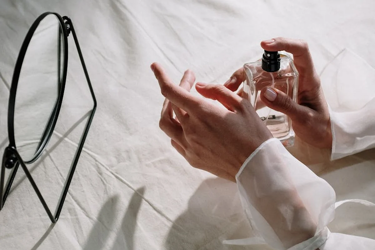 Hands in white sleeves holding a small spray bottle beside a round mirror on a white fabric surface.