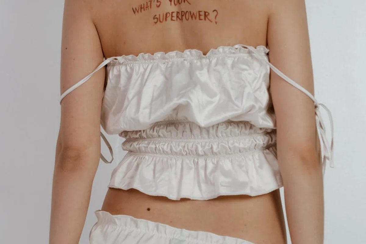 Back view of a person wearing a white ruched off-the-shoulder crop top; tattoo on the upper back reads 'WHAT'S YOUR SUPERPOWER?'