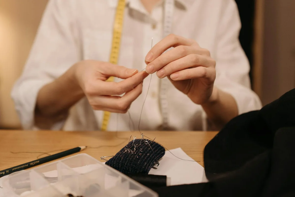 Hands threading a needle over a wooden table with fabric and a measuring tape in the background.