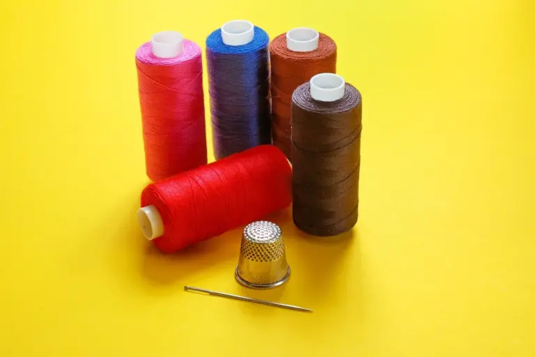 Assorted nylon thread spools in pink, red, blue, brown, and black arranged on a bright yellow background with a metal thimble and a needle.