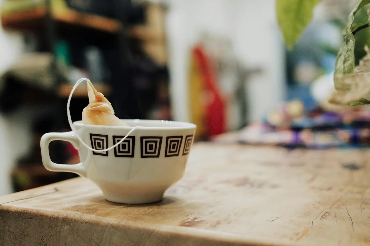 White ceramic teacup with a hanging tea bag on a wooden table in a cozy setting; blurred background shelves.
