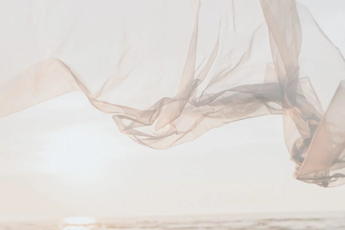 Beige sheer fabric blowing in the wind with a bright sun over a calm sea in the background.