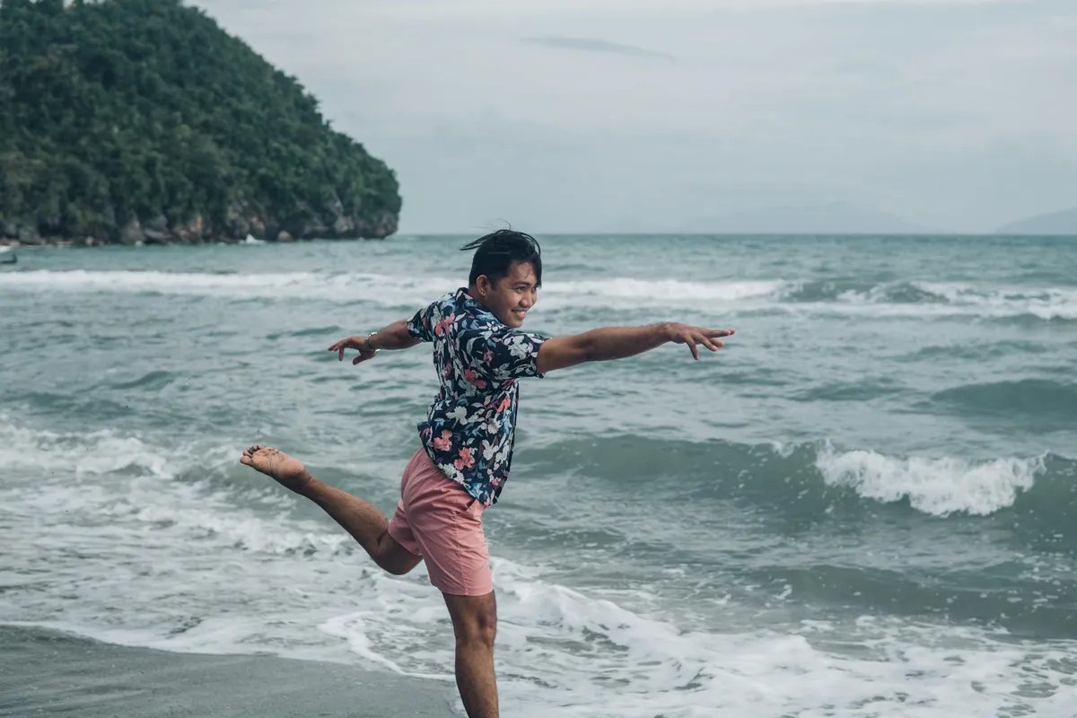 Person performing a dynamic balance on a sandy beach, wearing a floral shirt and pink shorts, with waves and a rocky headland in the background.
