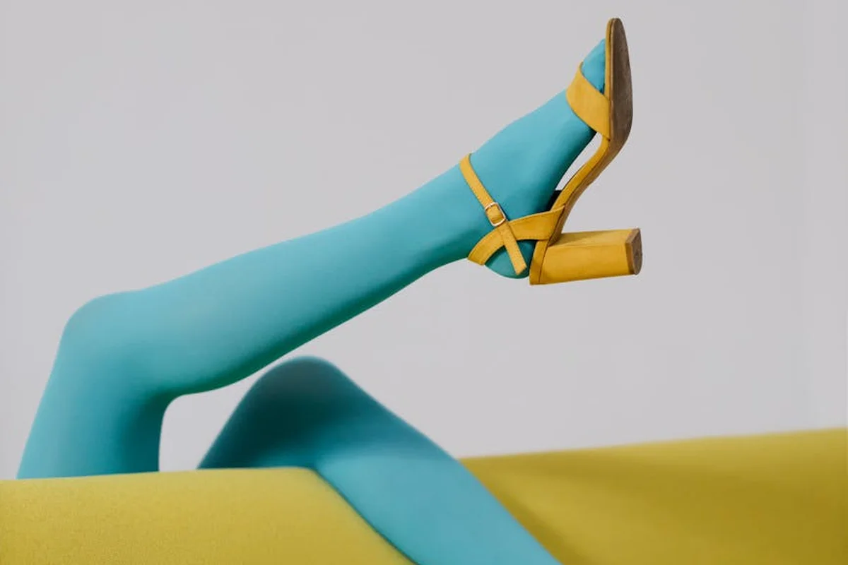Close-up of a teal nylon leg wearing bright yellow strap sandals, posed on a yellow surface.
