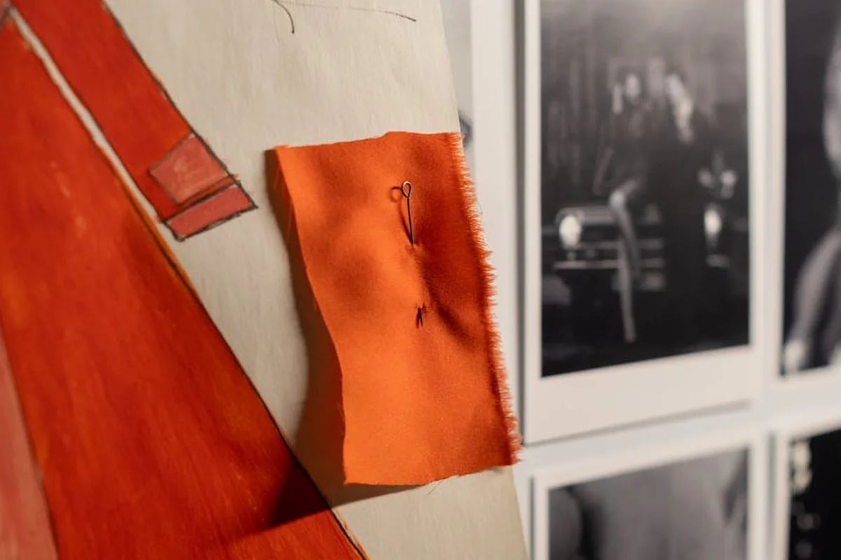 Close-up of a bright orange fabric swatch sewn and pinned to a wall, with blurred black-and-white photo frames in the background.