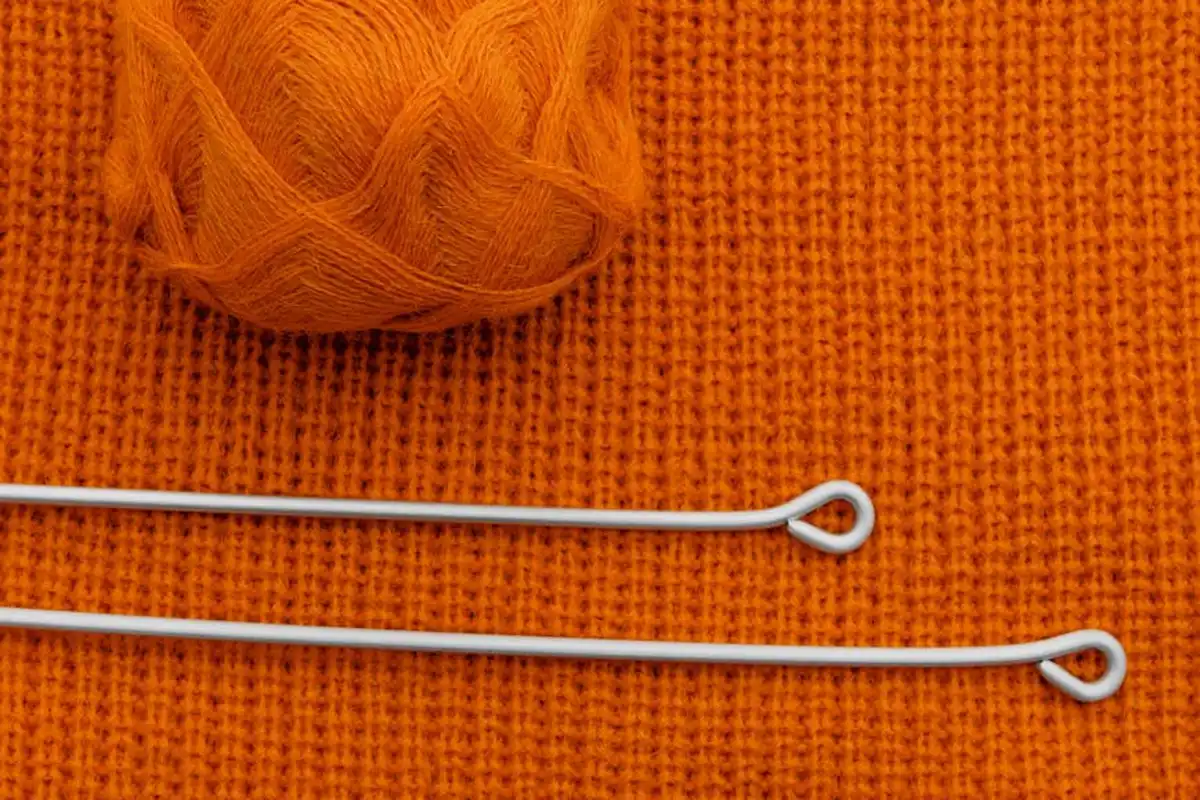 Close-up of bright orange knit fabric with a ball of yarn and two white looped stitch markers laid across the surface.
