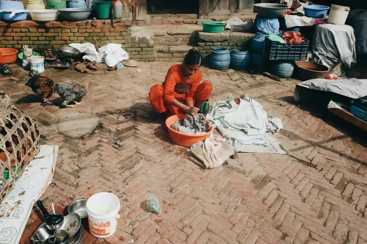 Person washing clothes by hand outdoors on a brick courtyard, with basins and laundry tools scattered around