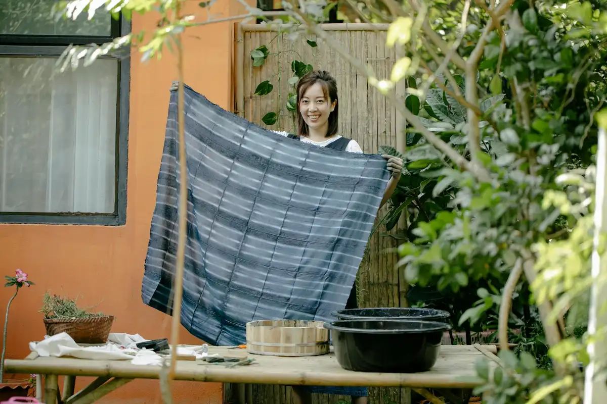 A person holds up a blue striped fabric outdoors, with dyeing supplies like a wooden drum and a black tub visible on a table, illustrating color selection and prep for dyeing Tencel and Lyocell.