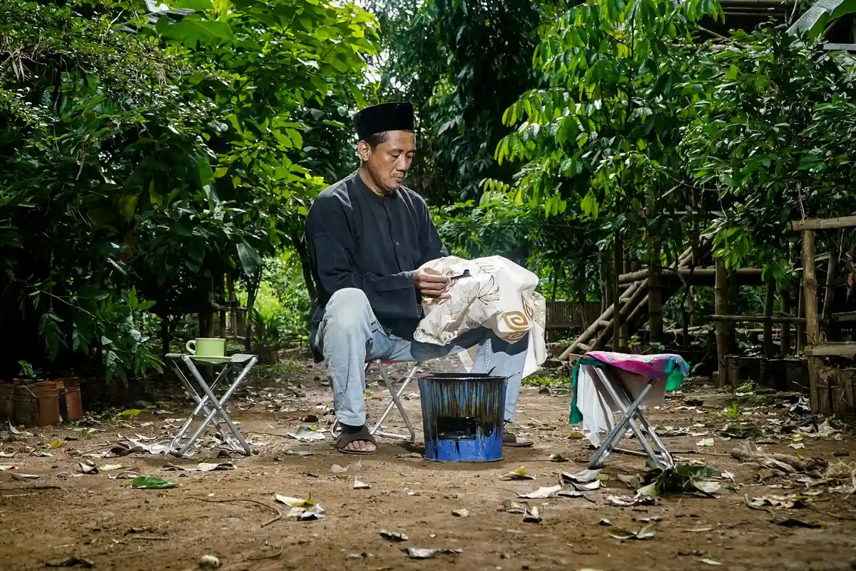A man wearing a dark shirt and cap sits outdoors, hand-washing a fabric over a small tub, surrounded by trees and drying racks.