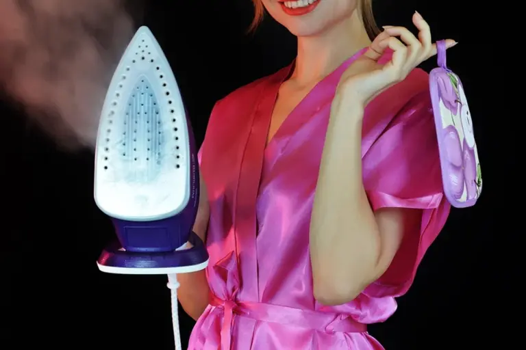 A person in a pink satin robe holds a blue and white steam iron with steam rising.