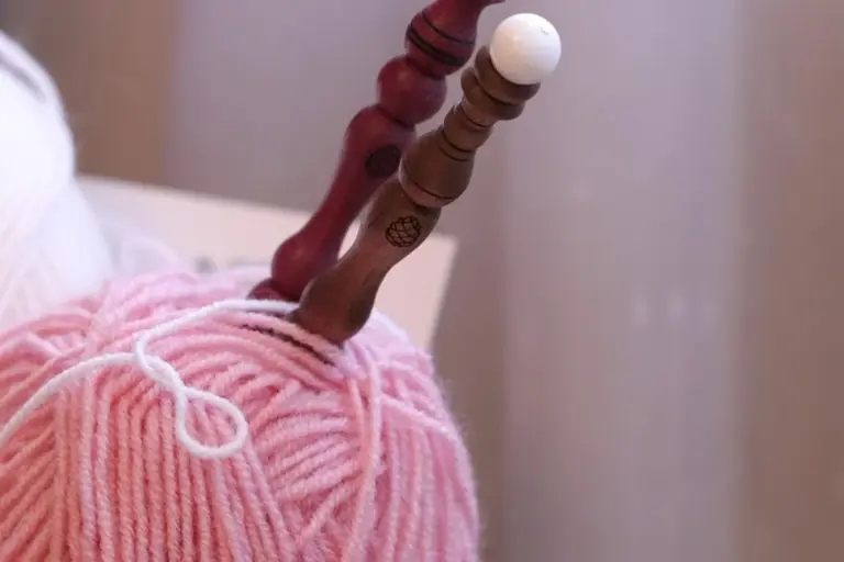 Close-up of pink wool yarn wrapped around knitting needles with a small white ball of yarn in the background.
