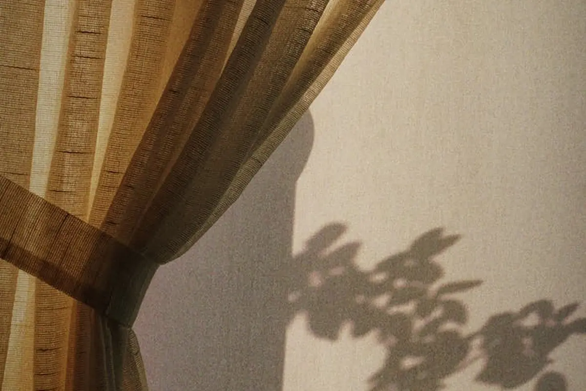 Beige polyester curtain with sunlight filtering through and leaf-shaped shadows on the wall