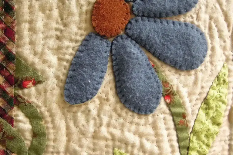 Close-up of a patchwork quilt with a blue fabric flower and orange center on a cream background, highlighting quilting stitches.