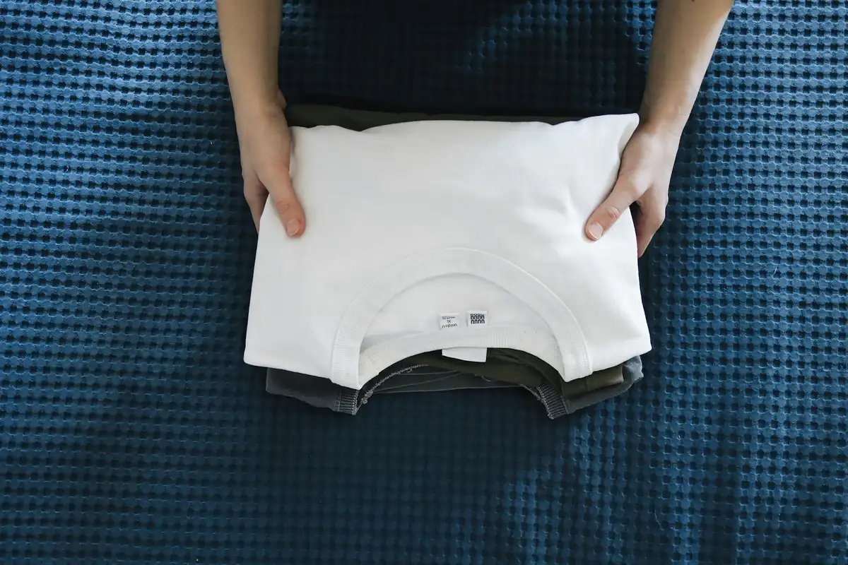 Person's hands fold a small stack of shirts on a blue textured surface.