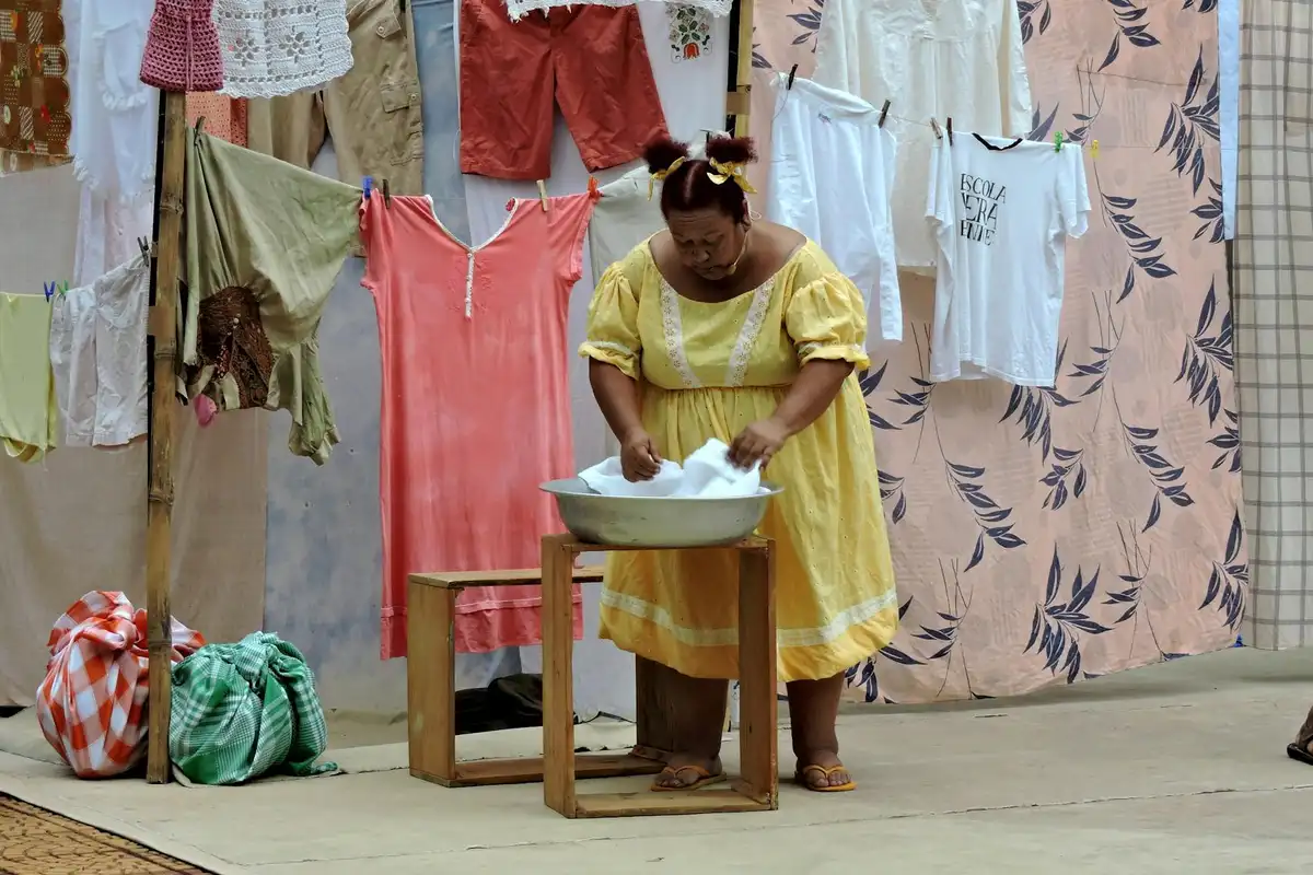 A woman in a yellow dress stands at a wooden basin washing clothes outdoors, with colorful garments hanging on lines behind her.