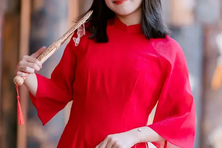 Person wearing a bright red traditional dress holds a decorative fan, shown from the chest up.