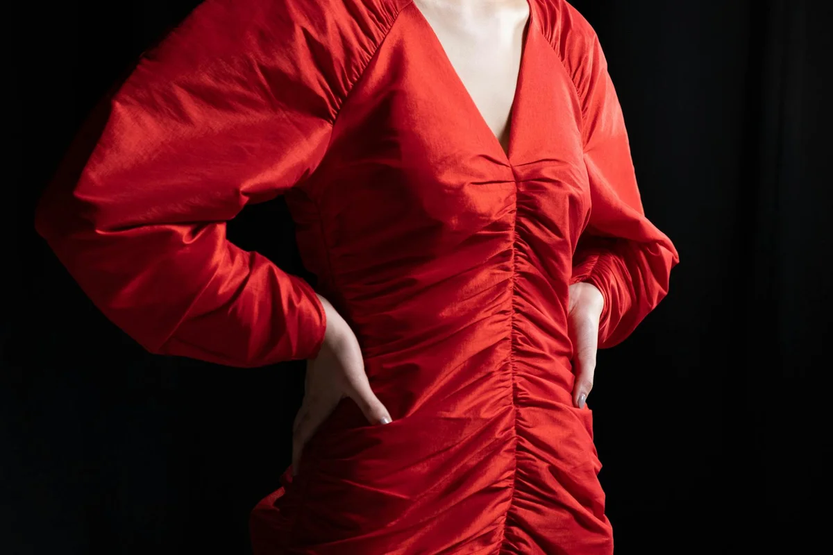 A person wearing a red silk ruched dress stands against a dark background.
