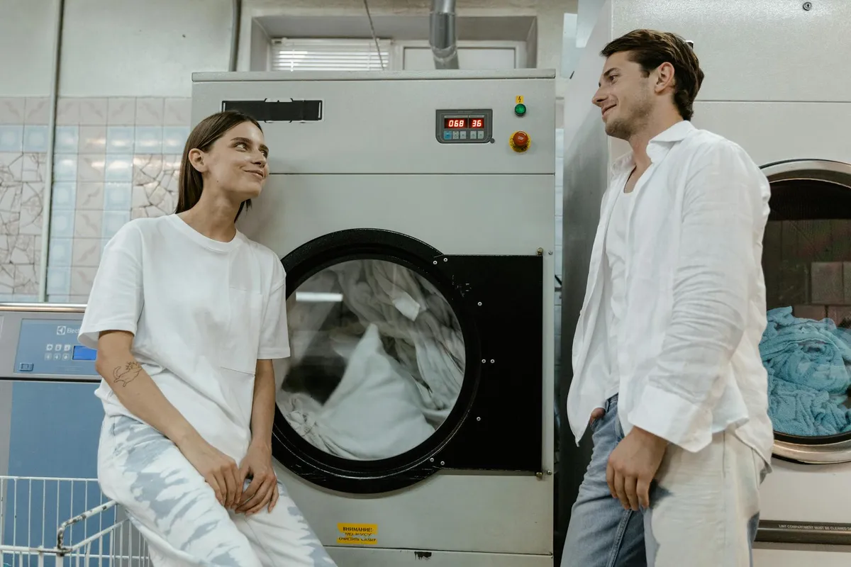 Two people stand near a front-loading washing machine in a laundromat, discussing the step-by-step care protocol for regenerated fabrics.