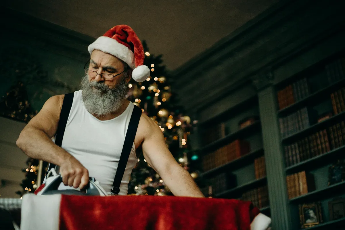 Person wearing a Santa hat irons a red fabric in a festive room with a Christmas tree and bookshelves.