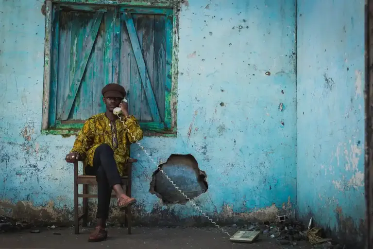 A person sits on a wooden chair against a weathered turquoise wall with a window and a large hole in the wall; they wear a yellow patterned shirt, dark pants, and a hat.
