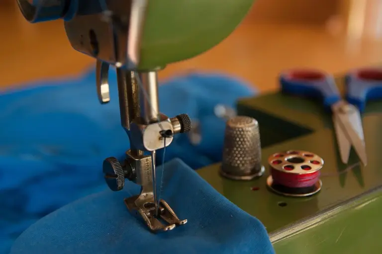 Close-up of a sewing machine needle and presser foot stitching blue fabric, with a spool and scissors in the background