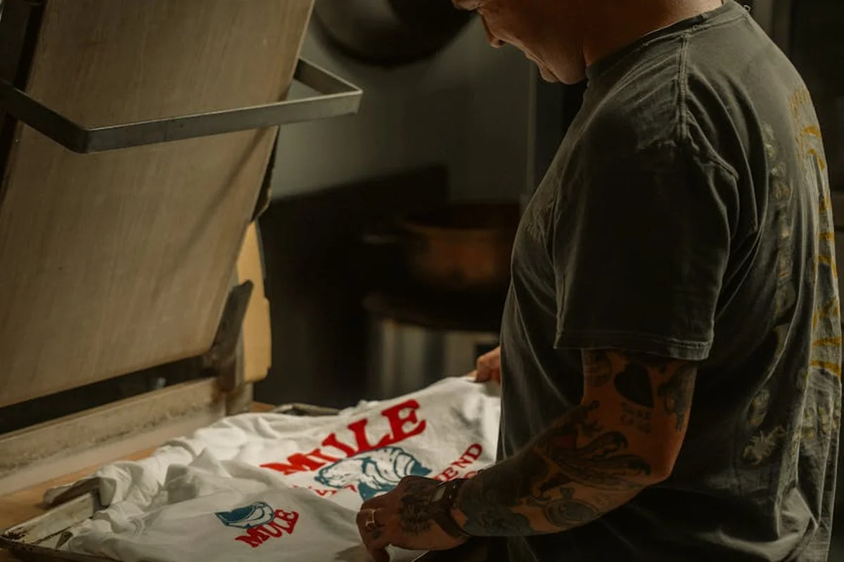 A worker operating a screen printing press, preparing a white T-shirt with red and blue graphics under the screen.