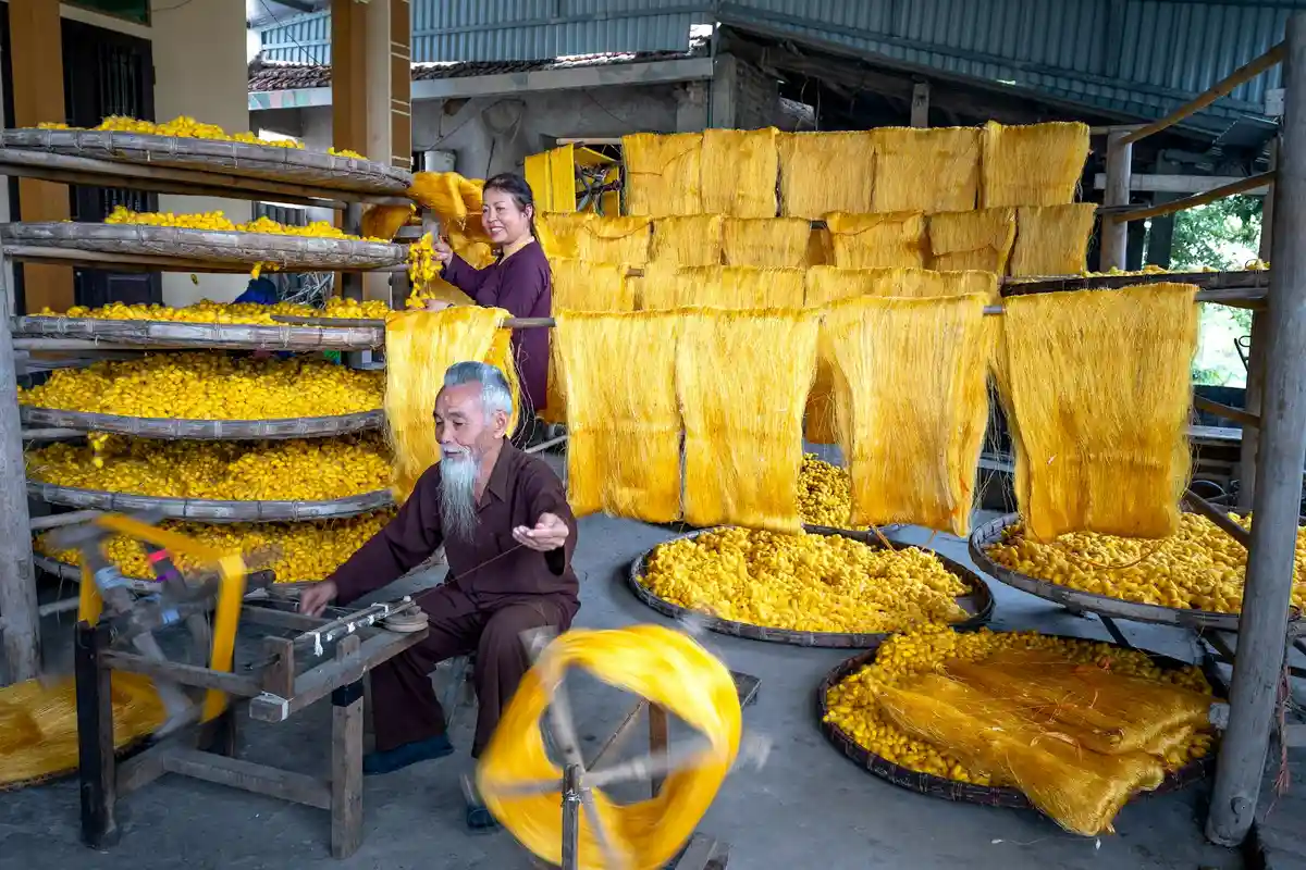 Two workers in a traditional silk workshop with bright yellow silk hanging to dry on racks; a reeling machine is visible in the foreground.