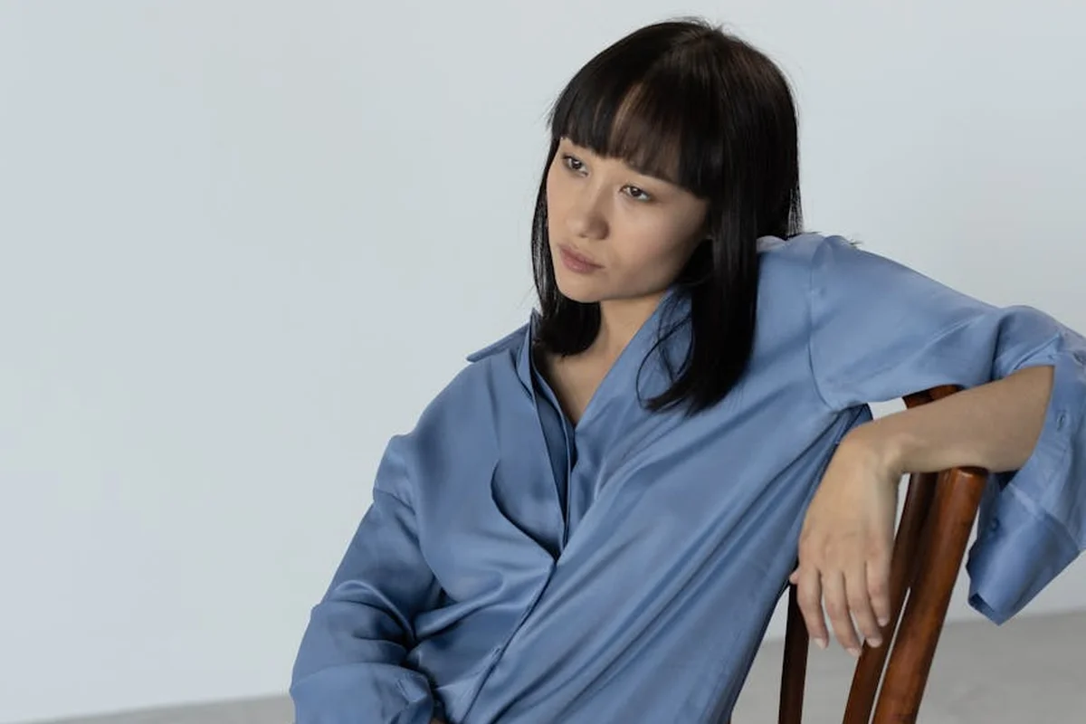 Person with dark hair wearing a blue silk blouse sits leaning on a wooden chair.
