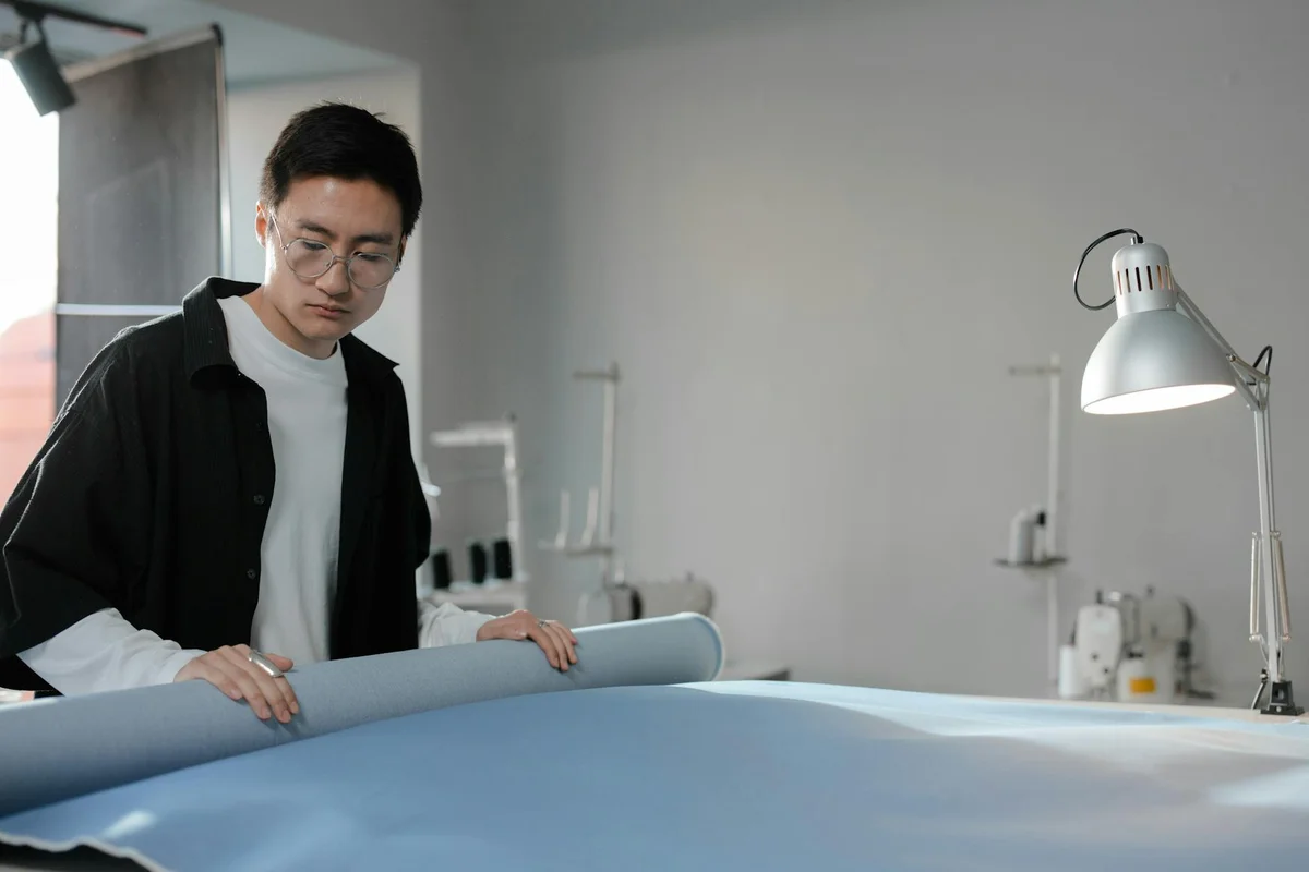 A designer unrolls and measures a wide sheet of light blue fabric in a bright studio, with a desk lamp in the background.