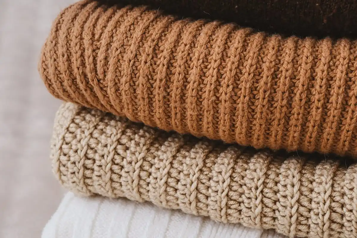 Stacked wool sweaters in warm brown, beige, and cream tones showing textured knit patterns.