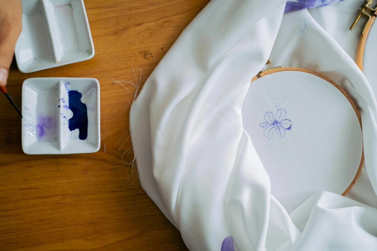 White fabric stretched in an embroidery hoop on a wooden table, with a small two-compartment paint palette and a splash of purple paint, illustrating the setup for painting on fabric to achieve a permanent finish.
