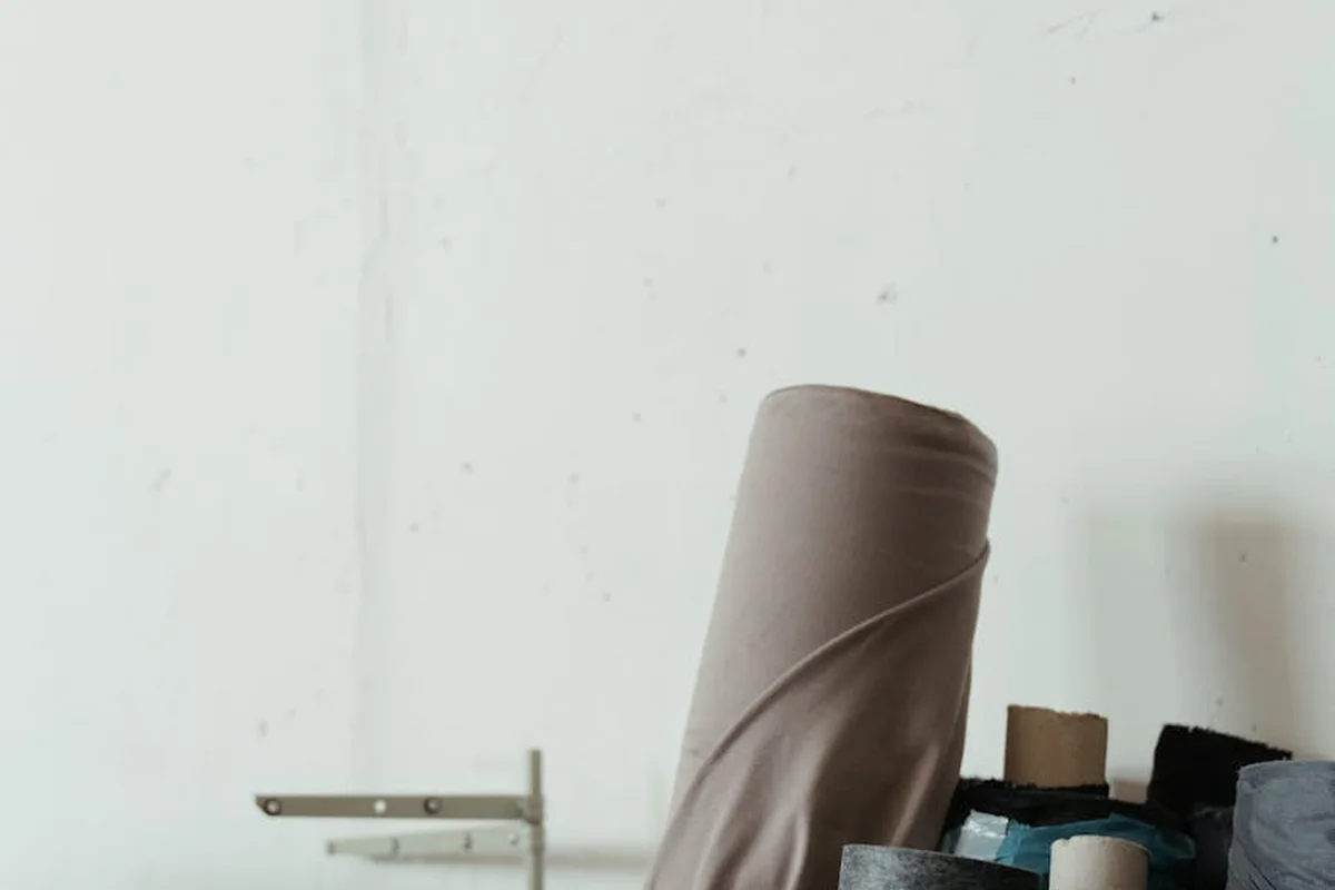 A light workspace featuring a rolled fabric leaning upright against a pale wall, with small sewing spools and materials nearby.