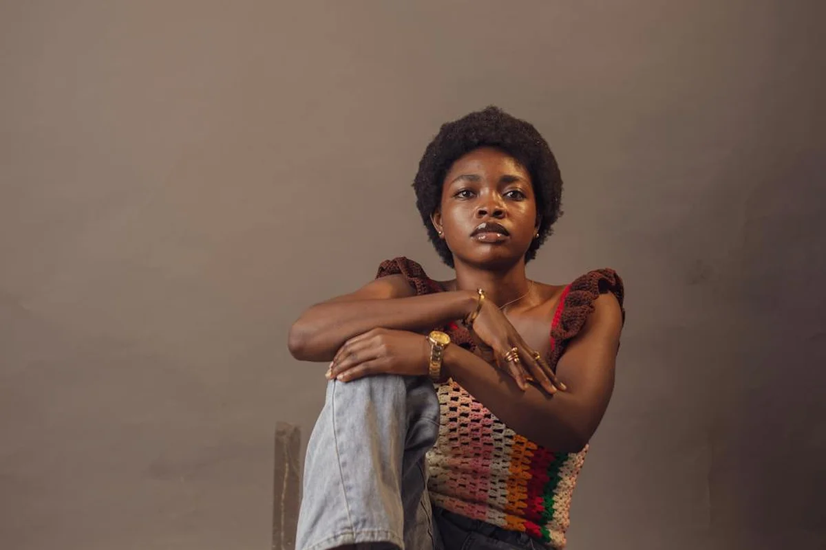 Portrait of a person with short natural hair wearing a colorful knitted top and jeans, sitting with one knee up against a plain brown backdrop.