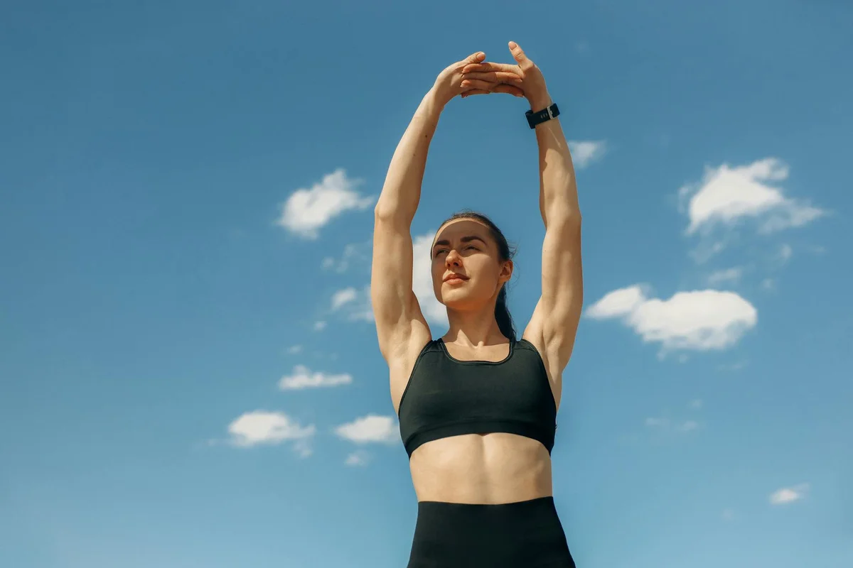 Person performing a standing stretch outdoors, arms raised overhead, wearing a black sports bra and leggings against a blue sky