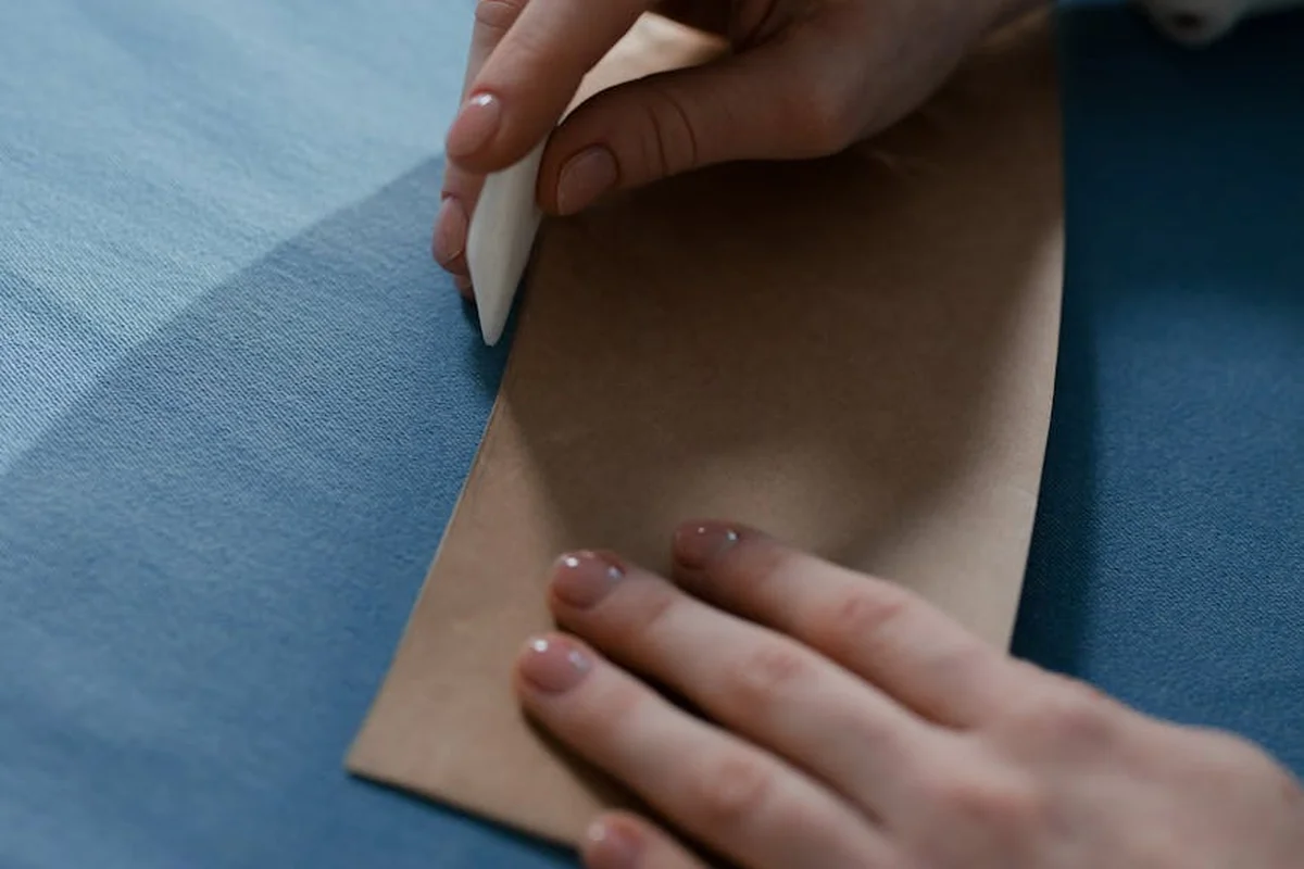 Close-up of hands guiding a piece of fabric with a small white pressing tool along the edge on a blue surface, illustrating seam preparation for stretch fabrics.