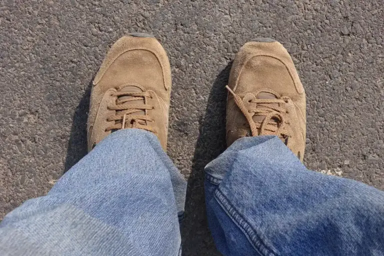 Top-down view of a person wearing tan suede shoes and blue jeans standing on a textured asphalt surface.