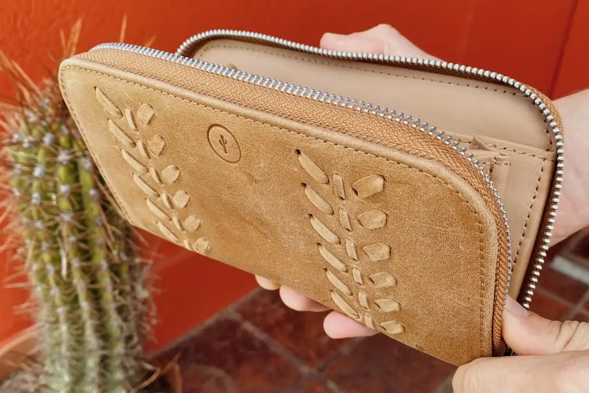 Close-up of a tan suede wallet being held by a hand, with a cactus and an orange wall in the background, highlighting suede texture.
