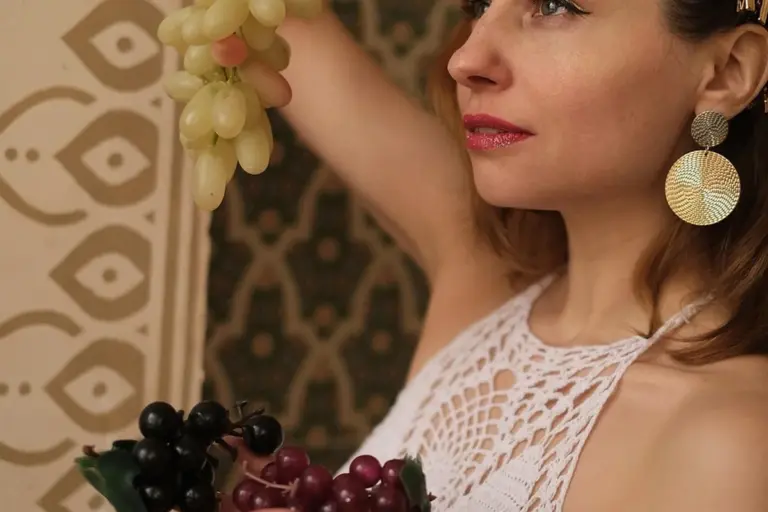 Portrait of a woman with a neutral expression wearing a white lace top and holding a bunch of grapes.