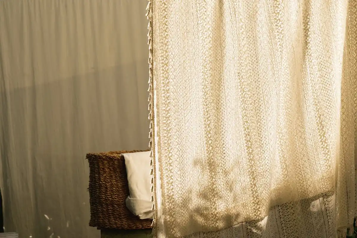 A wicker chair with a white cushion sits beside sheer, patterned curtains illuminated by warm light.