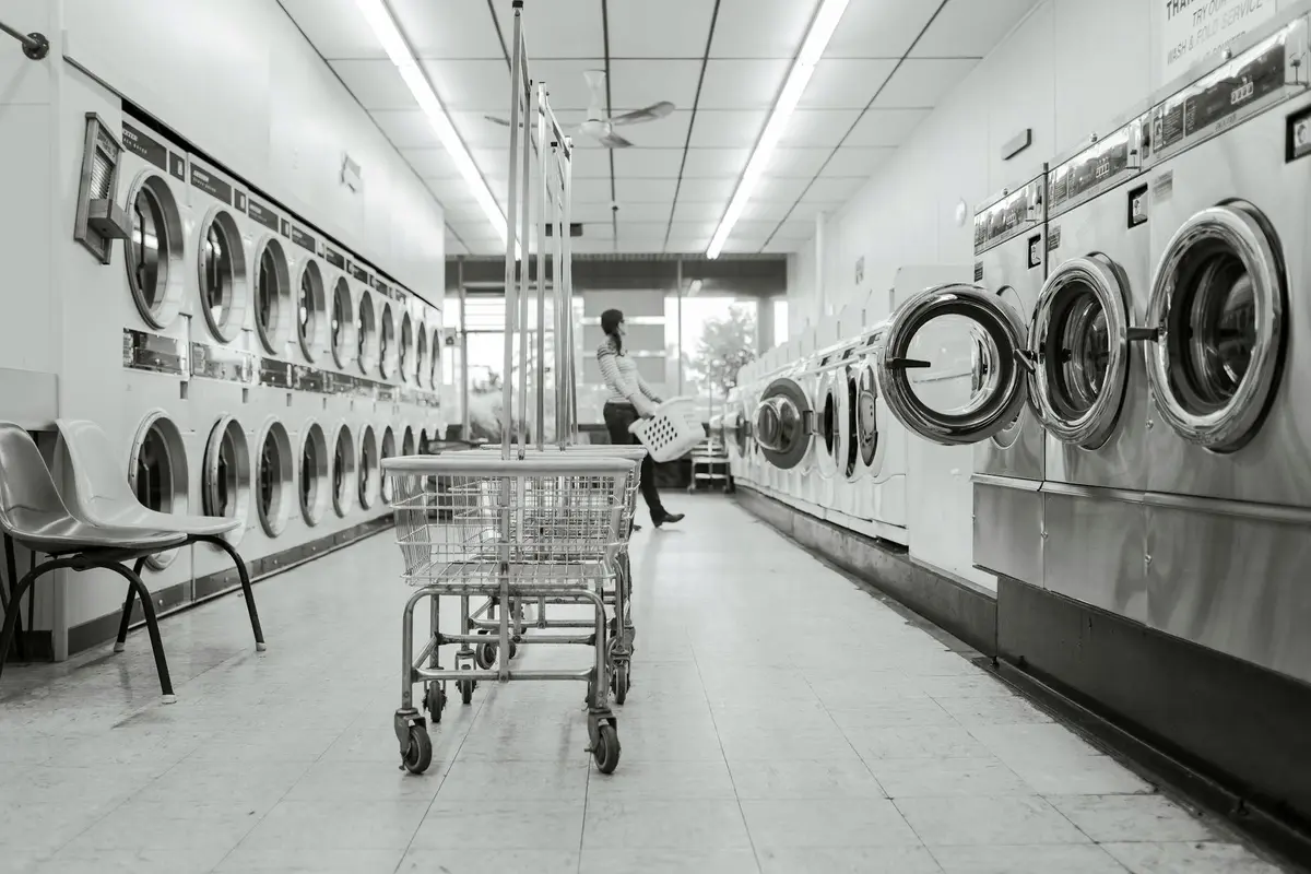 Black-and-white photo of a laundromat with row of front-loading washing machines on both sides and a shopper with a laundry cart in the center.